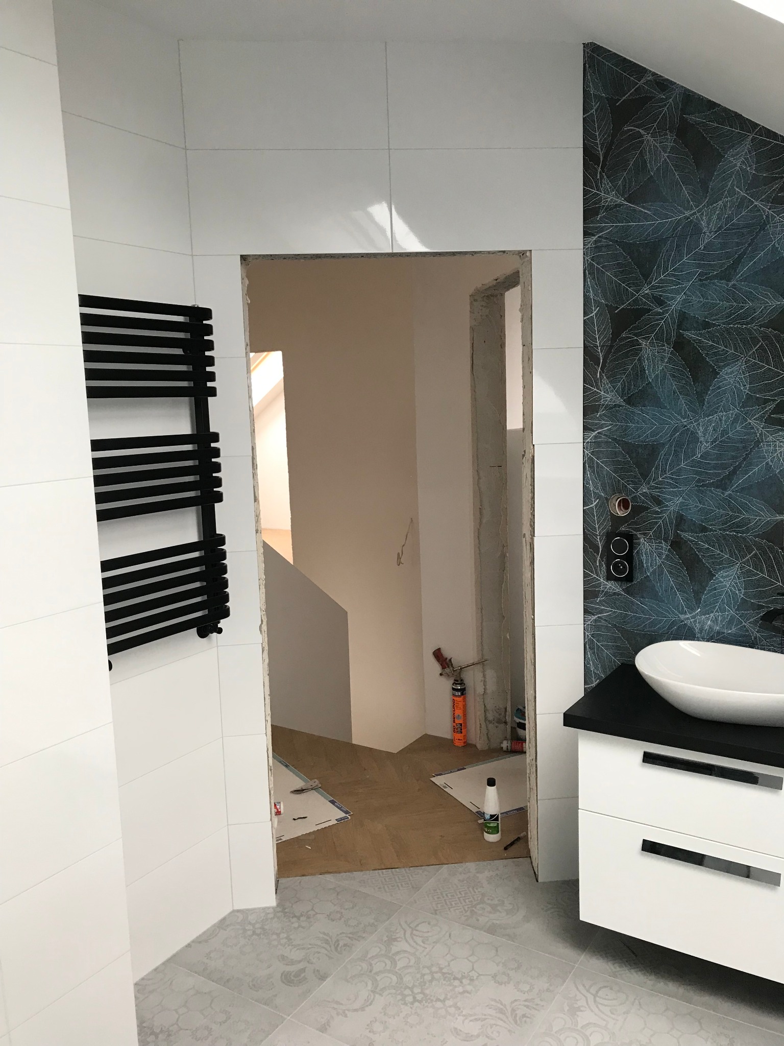 Remont łazienki w trakcie: położone białe płytki ścienne, dekoracyjna tapeta z motywem liści, grzejnik drabinkowy w kolorze czarnym, biała umywalka nablatowa, podłoga z szarymi płytkami we wzory...