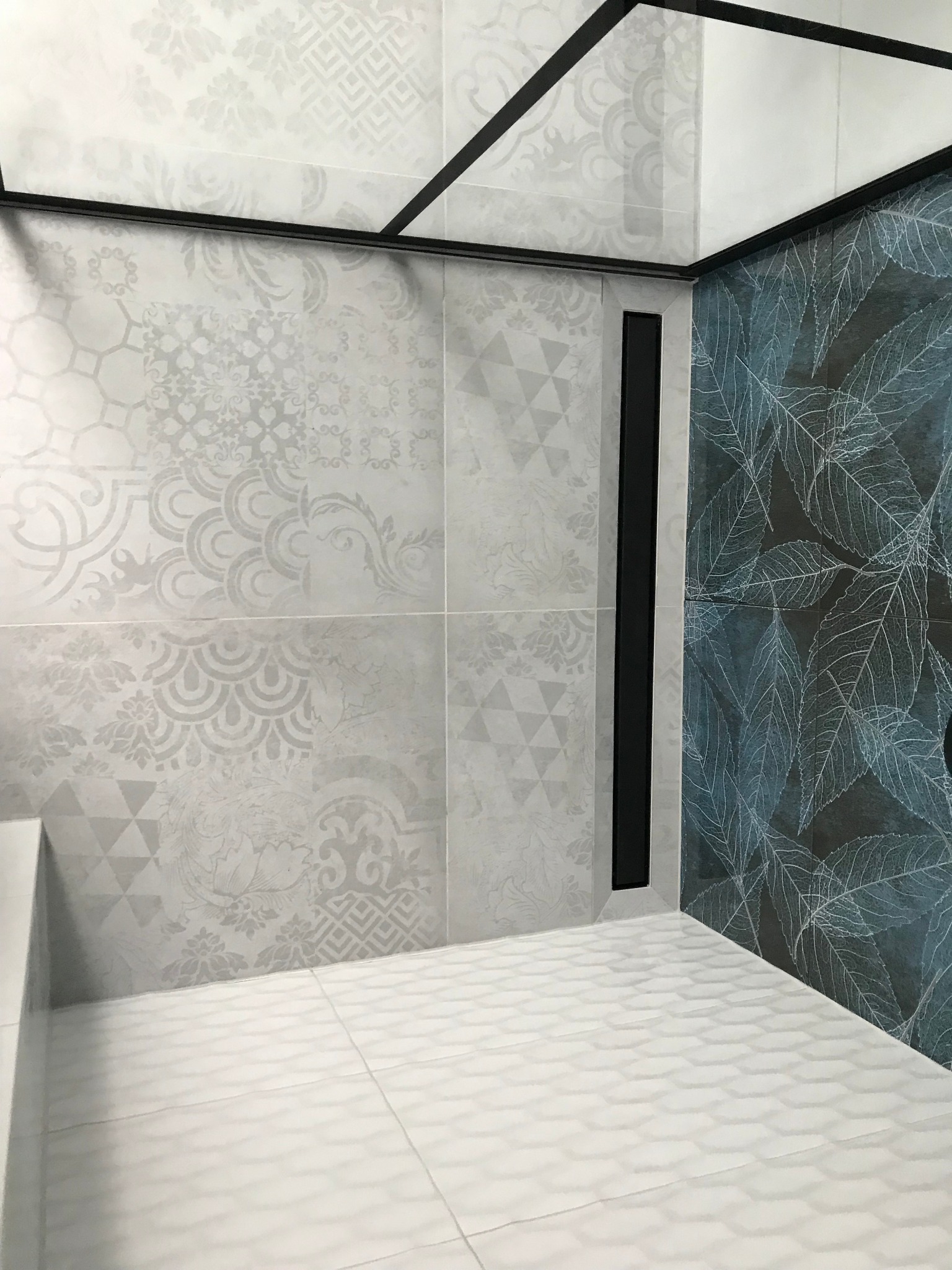 Wykończona kabina prysznicowa z różnymi wzorami płytek: szare płytki z ornamentami, ciemne z motywem liści i biała mozaika na podłodze, wykończona czarnymi profilami.