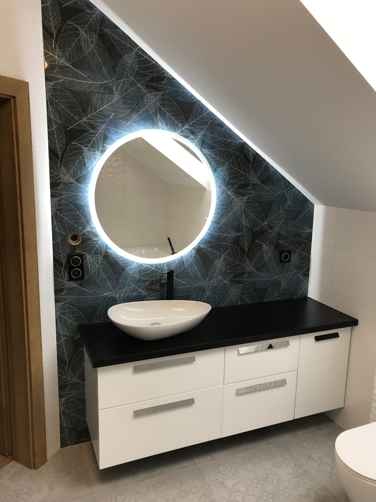 Nowoczesna łazienka na poddaszu z umywalką nablatową, okrągłym lustrem z podświetleniem LED, czarnym blatem i płytkami z motywem liści na ścianie.
