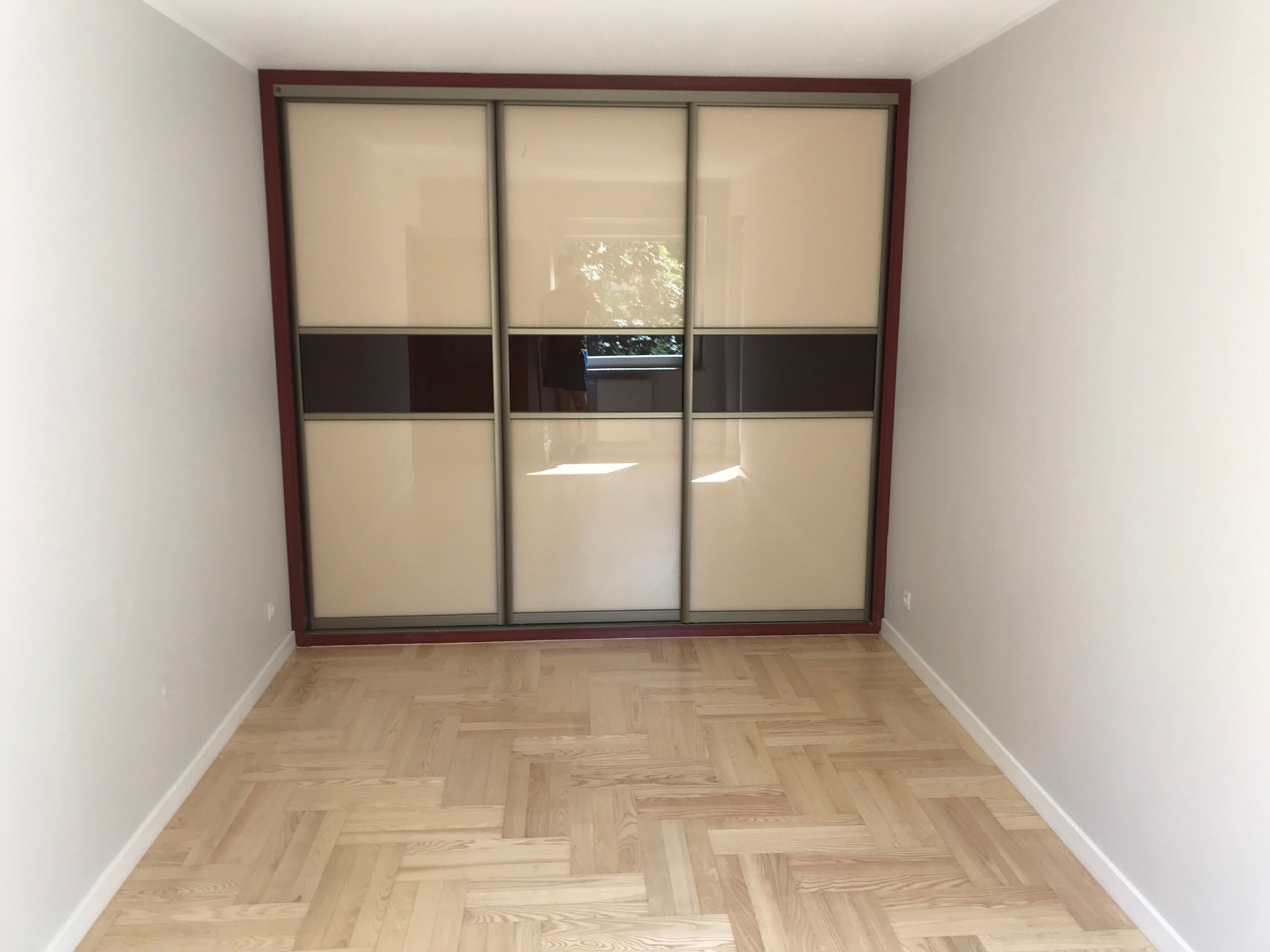 Pusta przestrzeń z parkietem ułożonym w jodełkę i szafą przesuwną z beżowymi drzwiami z brązowymi pasami na środku, obramowaną ciemnoczerwoną ramą.