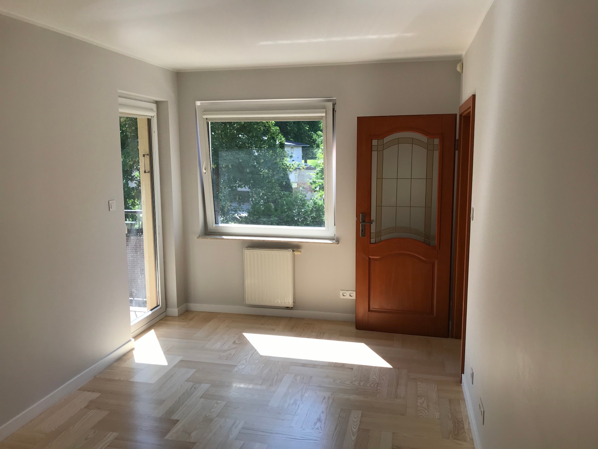 Jasne, puste pomieszczenie z parkietem w jodełkę, świeżo pomalowanymi szarymi ścianami, oknem z widokiem na zieleń i drewnianymi drzwiami z przeszkleniami.