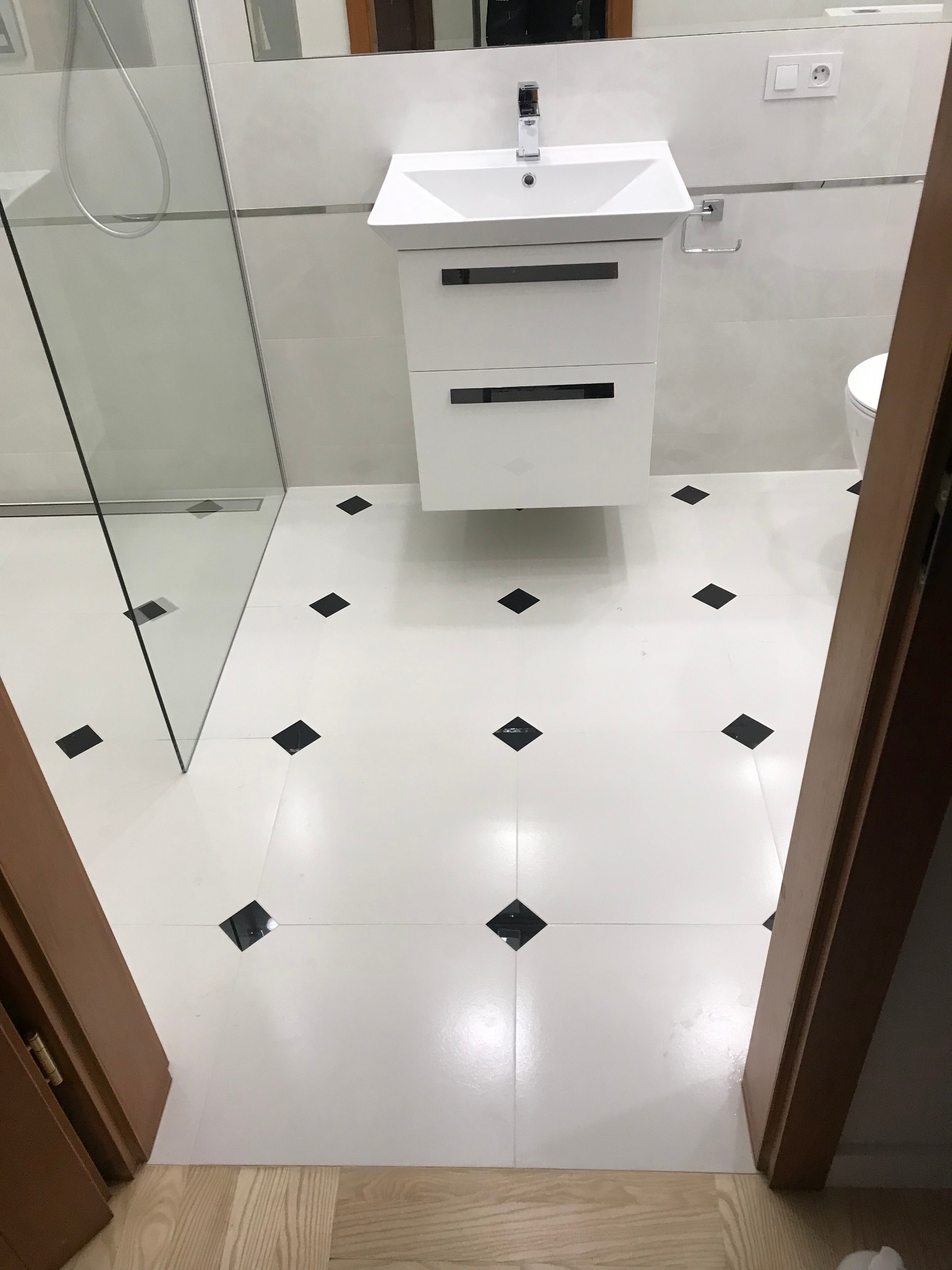 Nowoczesna łazienka z białymi płytkami podłogowymi z czarnymi akcentami, białą szafką pod umywalką i kabiną prysznicową ze szklaną ścianką, widziana zza drewnianej futryny drzwi.