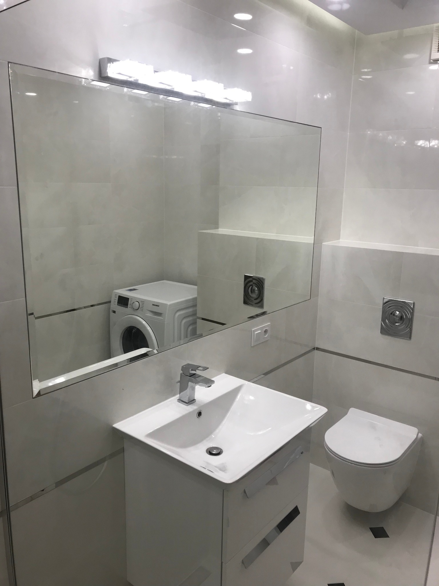 Nowoczesna, minimalistyczna łazienka z białymi płytkami, umywalką z szafką, podwieszaną toaletą i dużym lustrem, w którym odbija się pralka.