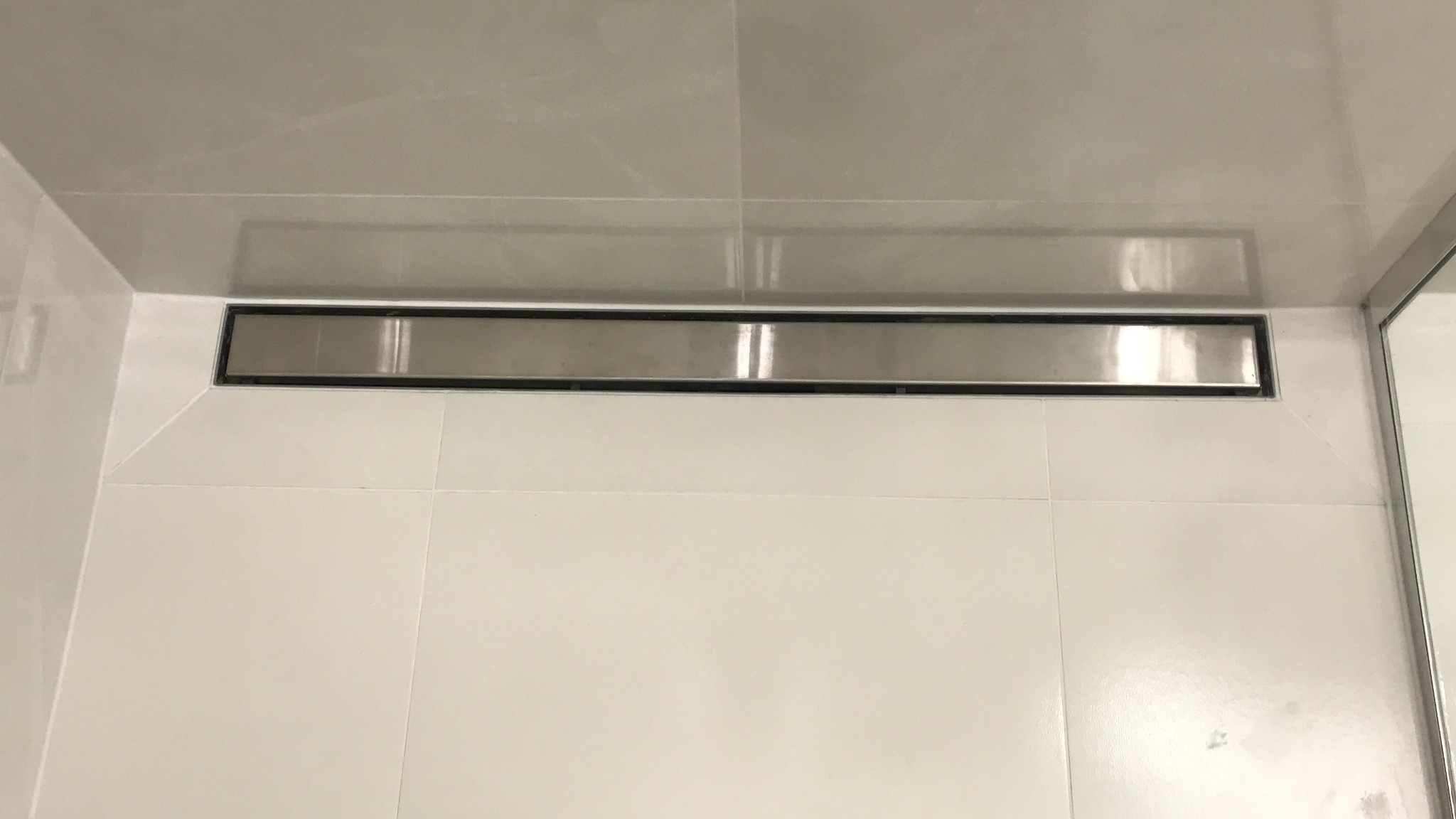 Szczegółowe ujęcie liniowego odpływu prysznicowego ze stali nierdzewnej, wkomponowanego w jasne, kwadratowe płytki ceramiczne, z widocznym odbiciem światła na metalowej powierzchni.