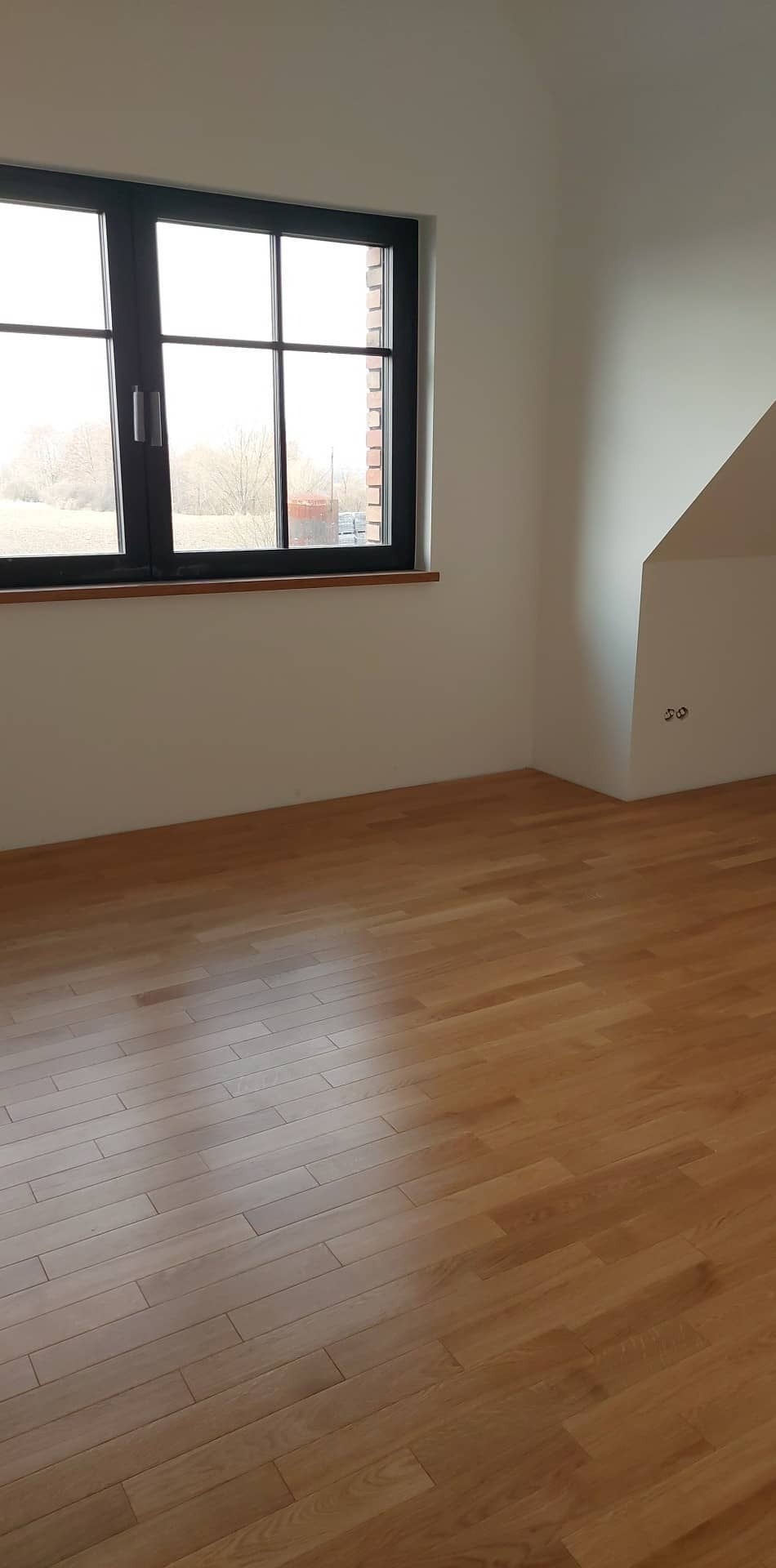 Pusty, jasny pokój z drewnianą podłogą, czarną ramą okienną i białymi ścianami, widoczne gniazdka elektryczne.