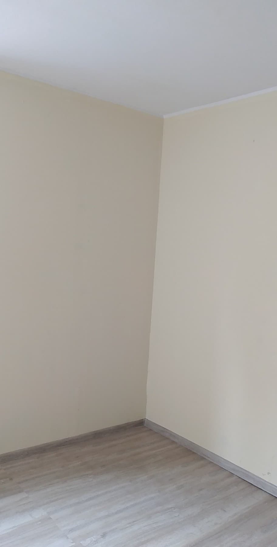 Pomalowany na kremowo róg pokoju z panelami podłogowymi, widoczna listwa przypodłogowa i sufitowa.