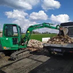 Zielona minikoparka Yanmar 825V załadowuje gruz do skrzyni białego samochodu dostawczego Tecnocar na tle błękitnego nieba z chmurami i zielonego pola.