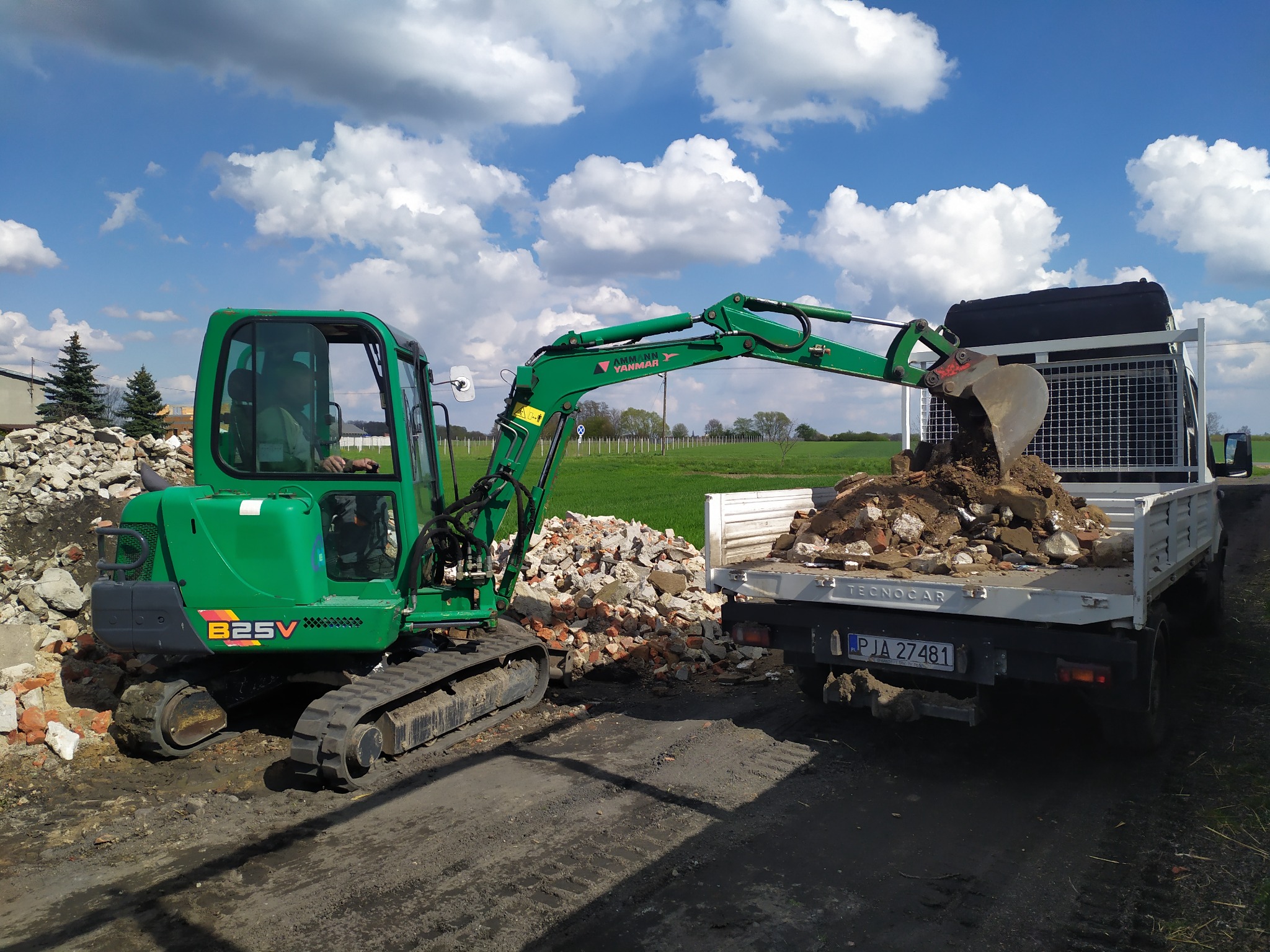 Zielona minikoparka Yanmar 825V załadowuje gruz do skrzyni białego samochodu dostawczego Tecnocar na tle błękitnego nieba z chmurami i zielonego pola.