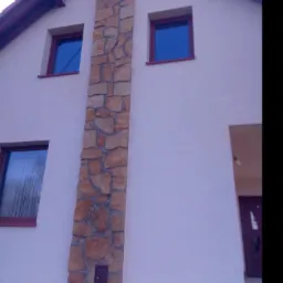 Fragment elewacji domu z dekoracyjnym pasem z kamienia naturalnego, okna z bordowymi ramami i widoczne elementy dachu.