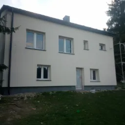 Dwukondygnacyjny dom jednorodzinny z jasną, świeżo pomalowaną elewacją, widoczne okna z białymi ramami, rynna, częściowo zakryte wejście z folią ochronną, trawnik w pierwszym planie, rusztowanie...