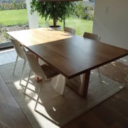 Jasny, drewniany stół z prostymi nogami, otoczony białymi krzesłami na tle dużego okna z widokiem na ogród, całość oświetlona popołudniowym słońcem tworzącym cienie na dywanie i podłodze.