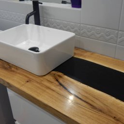 Techwood Przemysław Piątkowski - Biała, prostokątna umywalka nablatowa z czarną armaturą, osadzona na blacie z jasnego drewna z wtopioną czarną żywicą, na tle białych płytek z delikatnym wzorem.