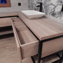 Techwood Przemysław Piątkowski - Łazienka z blatem drewnianym, umywalką nablatową i otwartą szufladą z przegródkami, czarna armatura i wzorzysta tapeta w tle.
