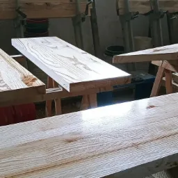 Surowe deski z jasnego drewna na stojakach w warsztacie, widoczne słoje i naturalna struktura drewna, przygotowane do dalszej obróbki.