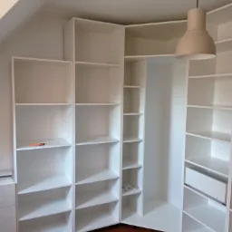 Biała szafa wnękowa z półkami, narożna, dopasowana do skosu dachu, z widocznym poziomem na podłodze i markerem na półce, oświetlona beżową lampą.