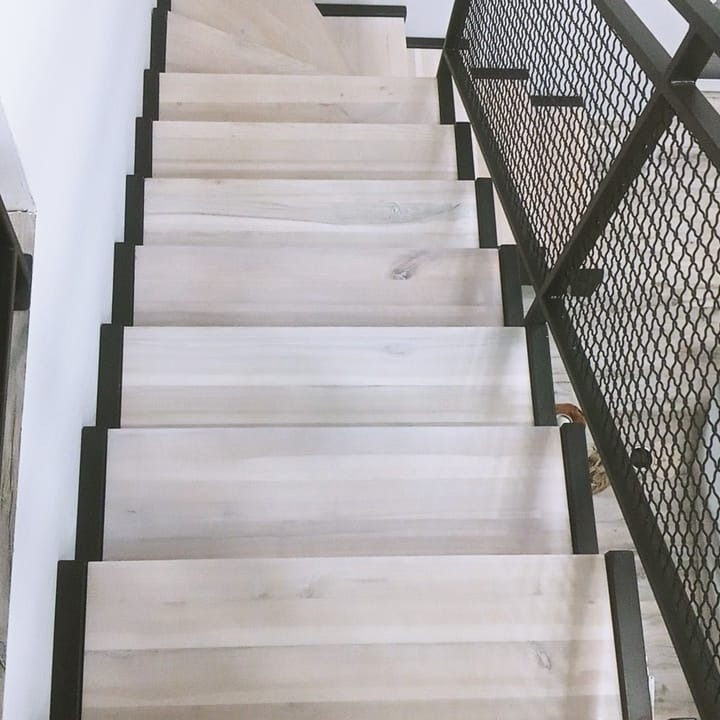 Jasne, drewniane stopnie z czarnymi krawędziami, widok z góry, czarna metalowa balustrada z siatką po prawej stronie, minimalistyczny design.