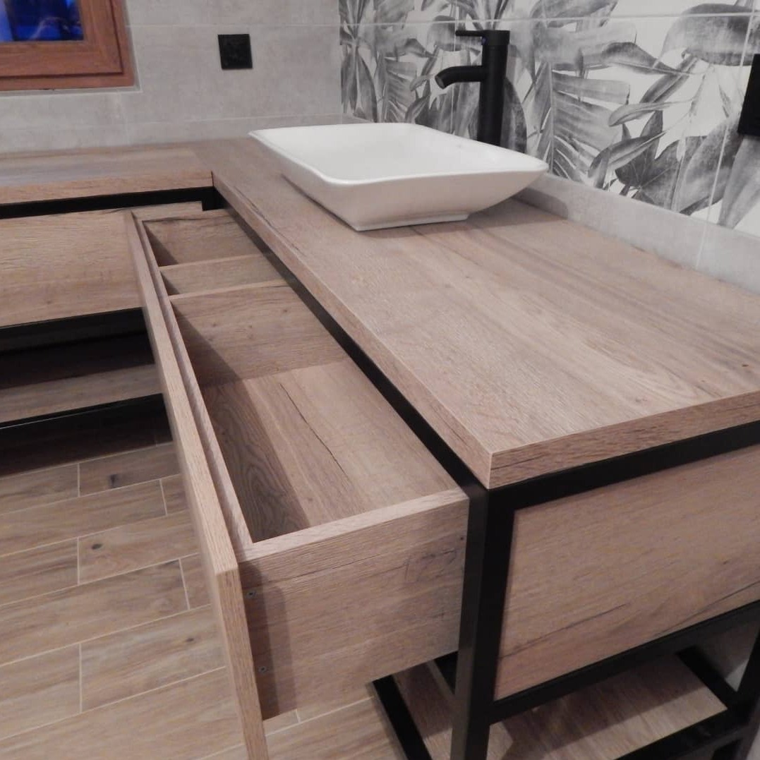 Łazienka z blatem drewnianym, umywalką nablatową i otwartą szufladą z przegródkami, czarna armatura i wzorzysta tapeta w tle.