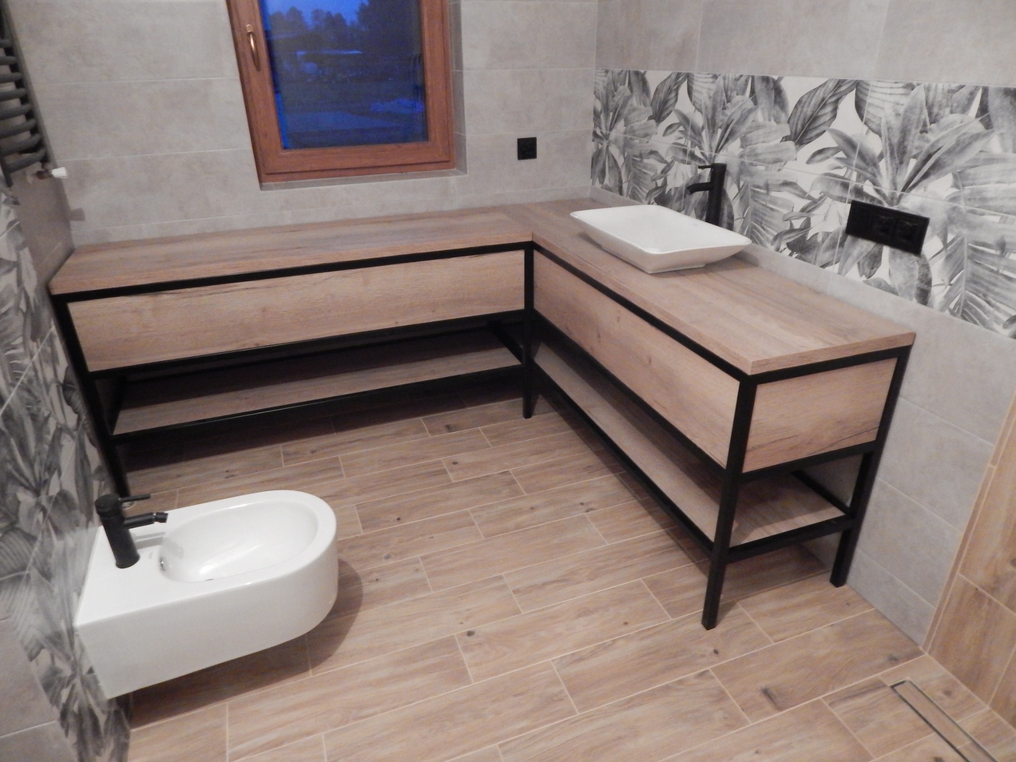 Łazienka z drewnianym blatem pod umywalkę i bidetem, czarne metalowe ramy, płytki imitujące drewno na podłodze, ściana z motywem liści.