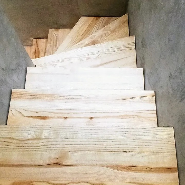 Widok z góry na jasne, drewniane stopnie schodów w surowym, betonowym wnętrzu, ukazujące naturalny rysunek słojów drewna.