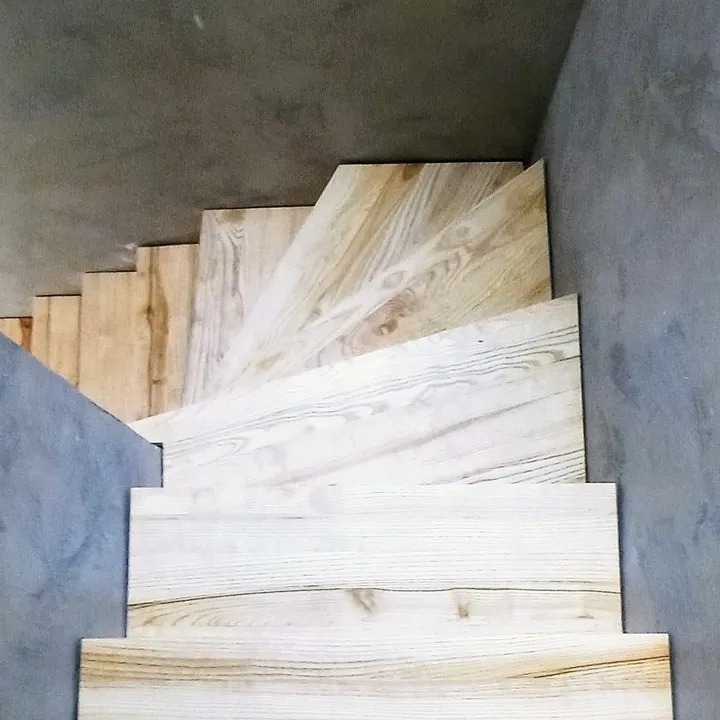 Wnętrze z betonowymi ścianami i schodami wyłożonymi jasnym drewnem z wyraźnym usłojeniem, widok z góry na dół.