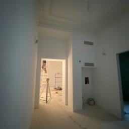 Usługi ogólnobudowlane STANEX - Prace malarskie w trakcie realizacji w Rząśniku: malarz na drabinie w jasnym wnętrzu, widoczne przygotowania i narzędzia malarskie.