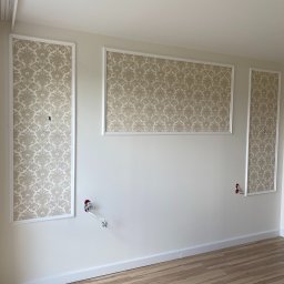 Usługi ogólnobudowlane STANEX - Pokój z panelami ściennymi z wzorzystą tapetą w białych ramach, widoczne gniazdka elektryczne z wystającymi przewodami, jasne panele podłogowe.