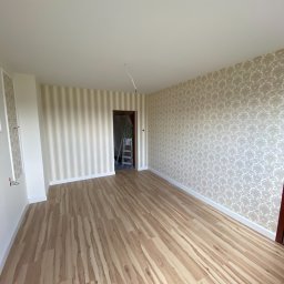 Usługi ogólnobudowlane STANEX - Pokój po remoncie z drewnianą podłogą, ścianami w paski i wzorzystą tapetą, widoczna drabina w przejściu do innego pomieszczenia.