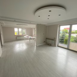 Usługi ogólnobudowlane STANEX - Jasne, przestronne wnętrze z parkietem w odcieniach szarości, okrągłym plafonem z oświetleniem punktowym i dużymi oknami balkonowymi z widokiem na zieleń.