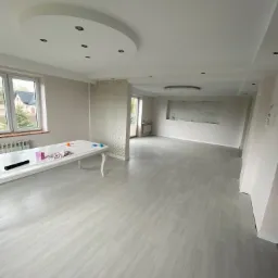 Jasne panele podłogowe w przestronnym, odnowionym salonie z białym stołem malarskim, dekoracyjną ścianą i oświetleniem sufitowym.