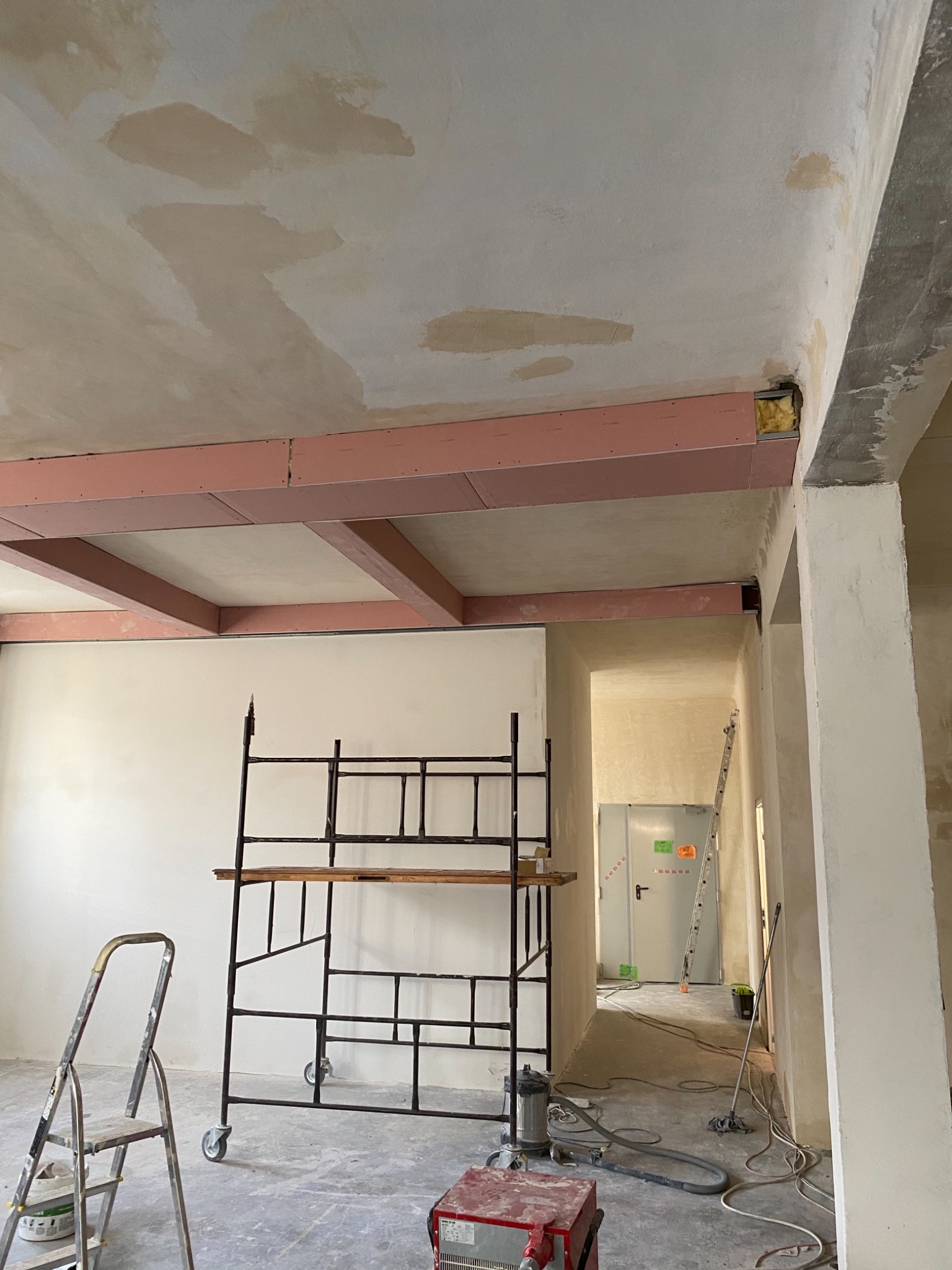 Wnętrze w trakcie prac wykończeniowych: widoczne rusztowanie, drabina, niezabezpieczone przewody, sufit z widocznymi śladami szpachlowania i konstrukcja z płyt kartonowo-gipsowych z różową izolacją...
