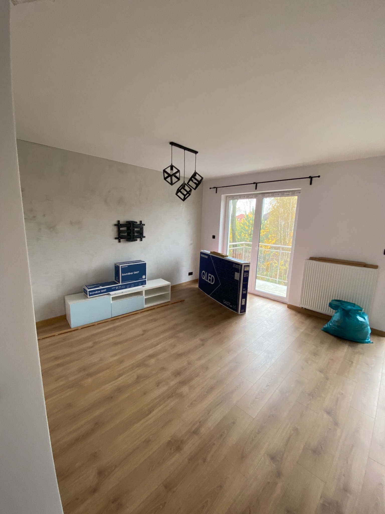 Pokój z panelami podłogowymi, świeżo pomalowaną ścianą z efektem betonu, zamontowanym uchwytem na telewizor, skrzynkami z nowym soundbarem i telewizorem QLED przed balkonem, oraz workiem na śmieci