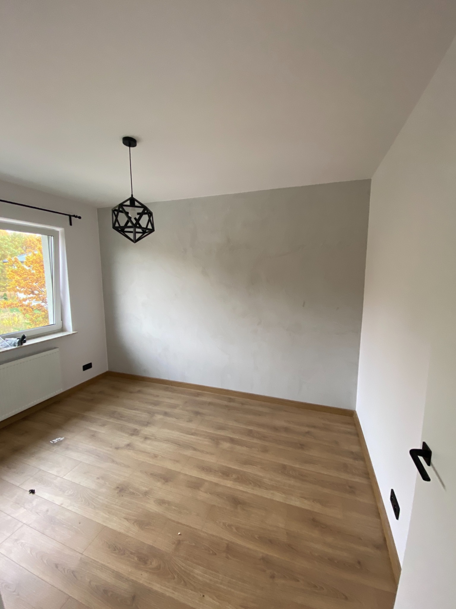 Pusty pokój po remoncie z panelami podłogowymi, ścianą z efektem betonu i geometryczną lampą wiszącą. Widok na jesienne drzewa za oknem.