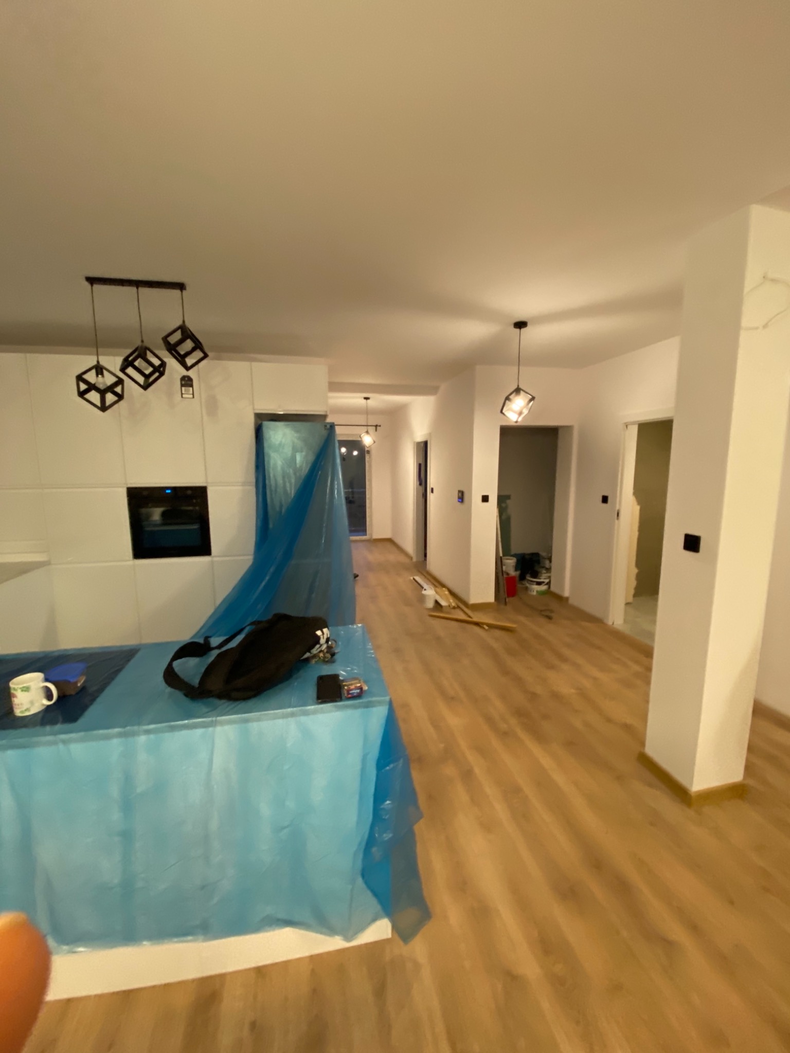 Wnętrze mieszkania w trakcie prac wykończeniowych: podłoga wyłożona panelami, ściany pomalowane na biało, meble kuchenne i sprzęty zabezpieczone folią ochronną, widoczne narzędzia i materiały...