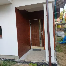 Dwukolorowa elewacja domu z brązowym akcentem imitującym drewno przy wejściu, brązowa rynna i częściowo rozstawione rusztowanie z boku.