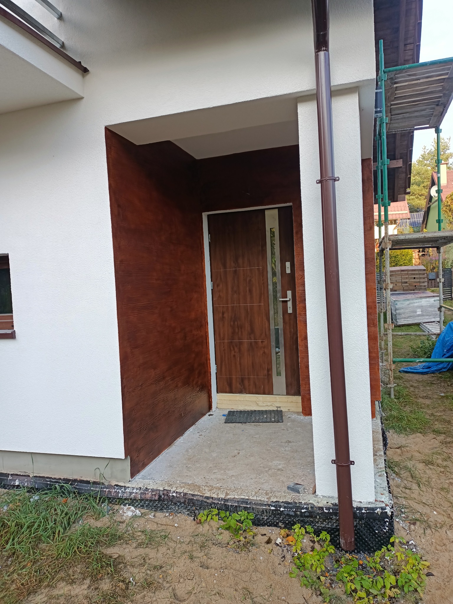 Dwukolorowa elewacja domu z brązowym akcentem imitującym drewno przy wejściu, brązowa rynna i częściowo rozstawione rusztowanie z boku.