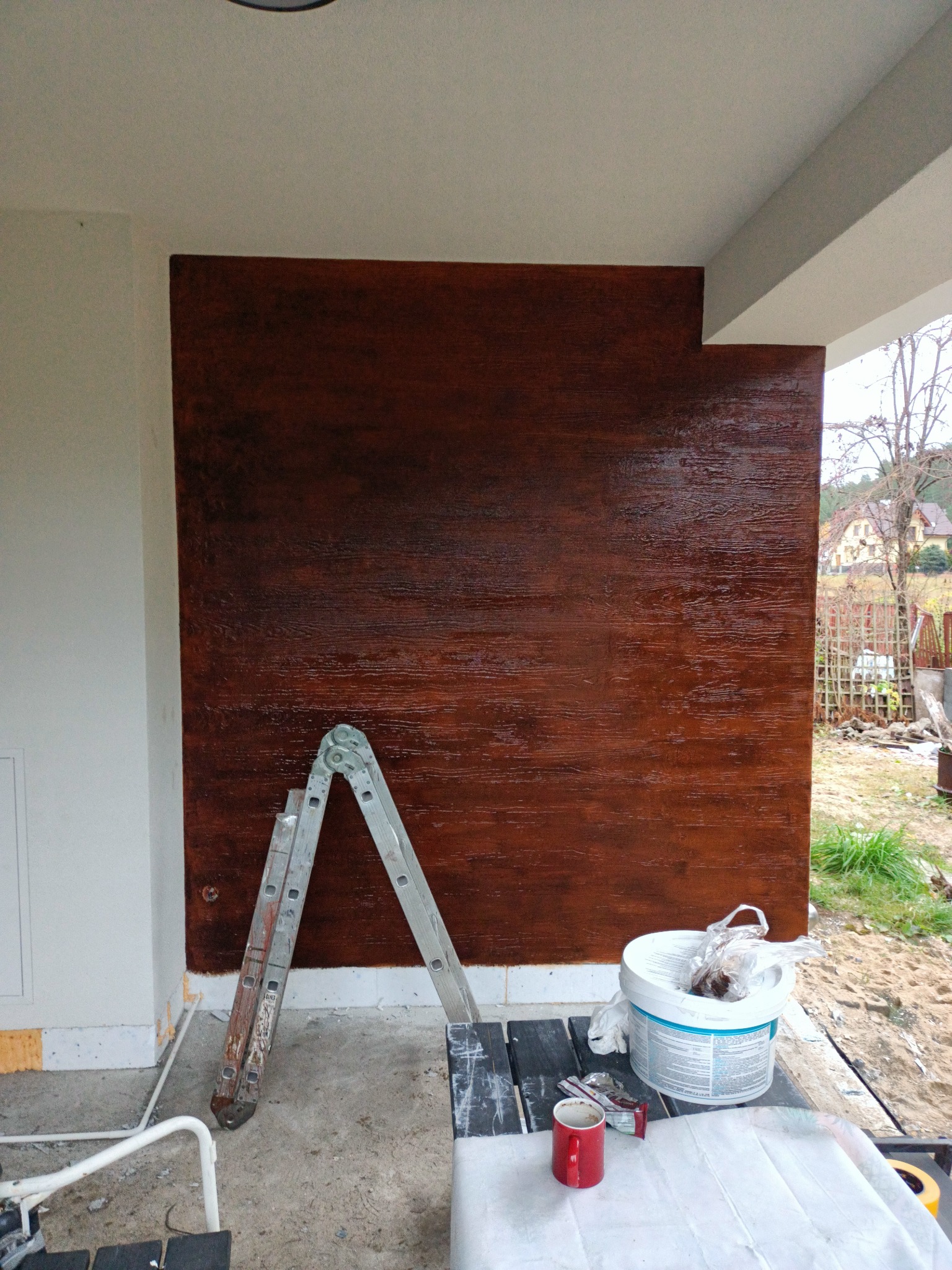 Wykonana elewacja zewnętrzna imitująca drewno, widoczna drabina, wiadro z farbą i kubek na stole, teren budowy.