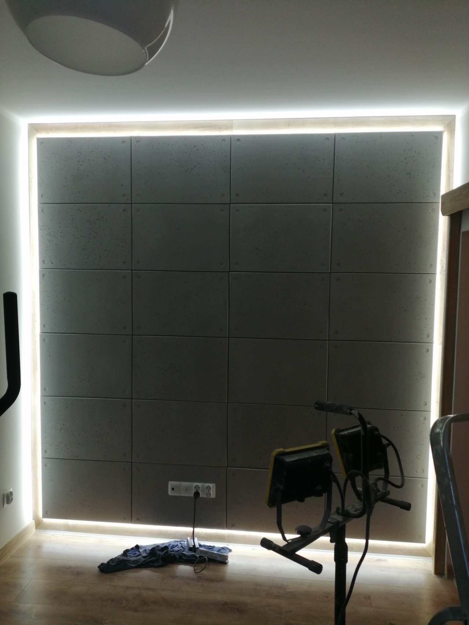 Ściana wyłożona kwadratowymi panelami z oświetleniem LED wzdłuż krawędzi, na podłodze leży niebieska szmatka i przedłużacz, a w kadrze widoczny jest statyw z dwoma lampami halogenowymi.