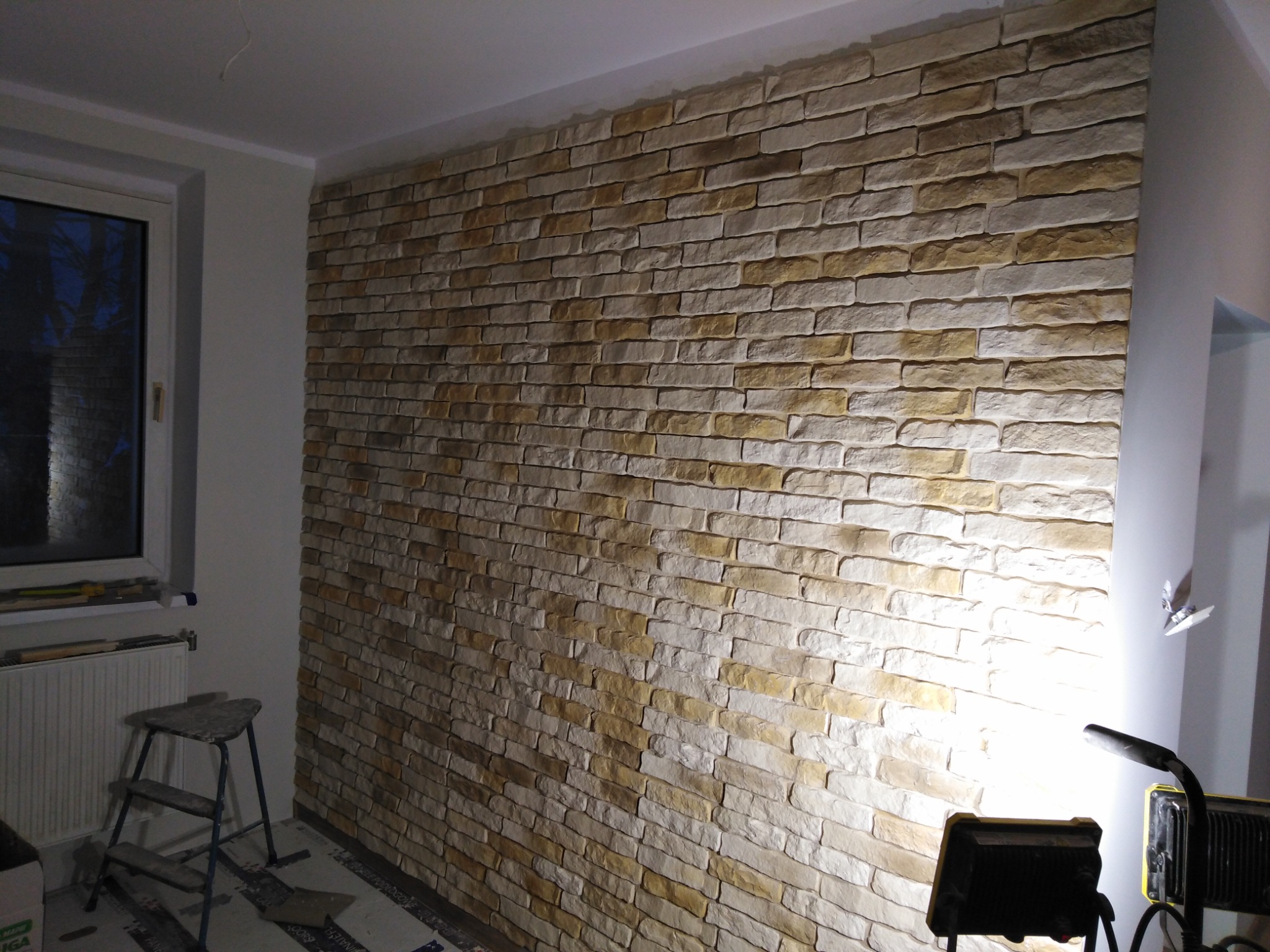 Ściana wewnętrzna wykończona dekoracyjną okładziną imitującą kamień w odcieniach beżu i brązu, oświetlona lampą budowlaną, widoczna drabinka malarska i grzejnik pod oknem.