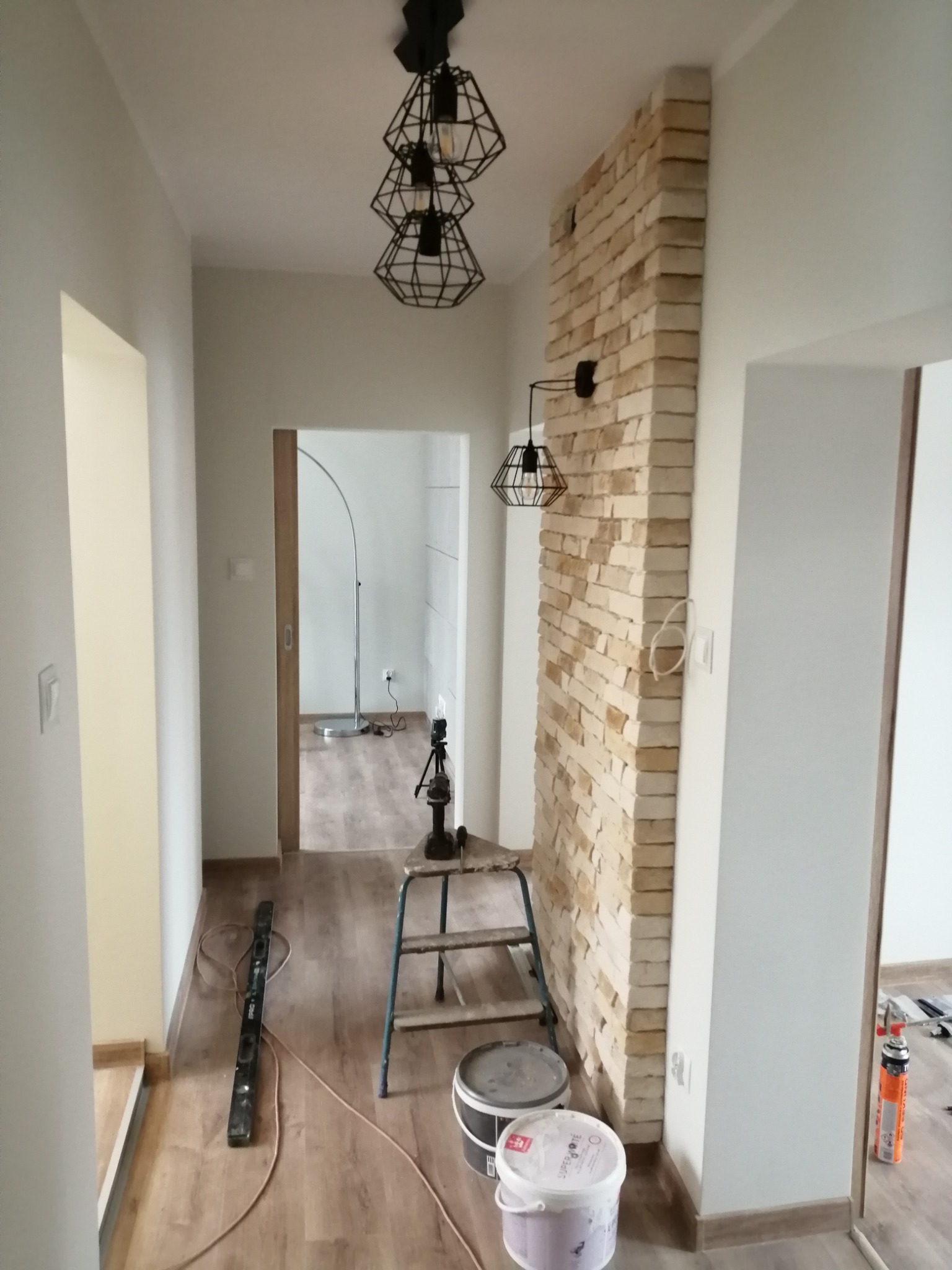 Korytarz w trakcie wykańczania z dekoracyjną ścianą z cegły, nowoczesne lampy wiszące, drabina, poziomica i puszki z farbą na podłodze.