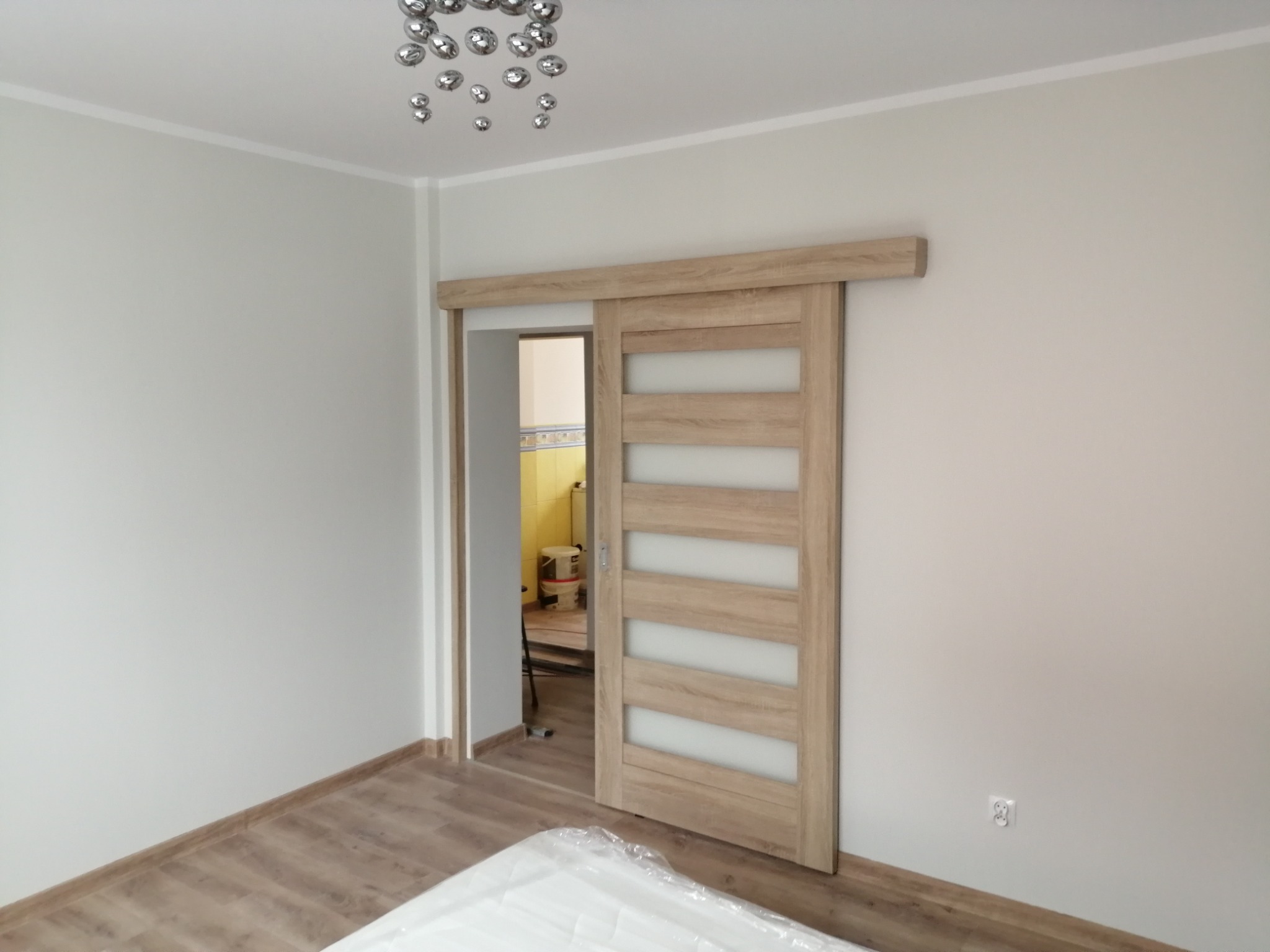 Zamontowane przesuwne drzwi drewniane z mlecznymi szybami, widok na odnowione wnętrze z żółtą ścianą i wiaderkiem farby w tle, na podłodze częściowo przykryta folią ochronną