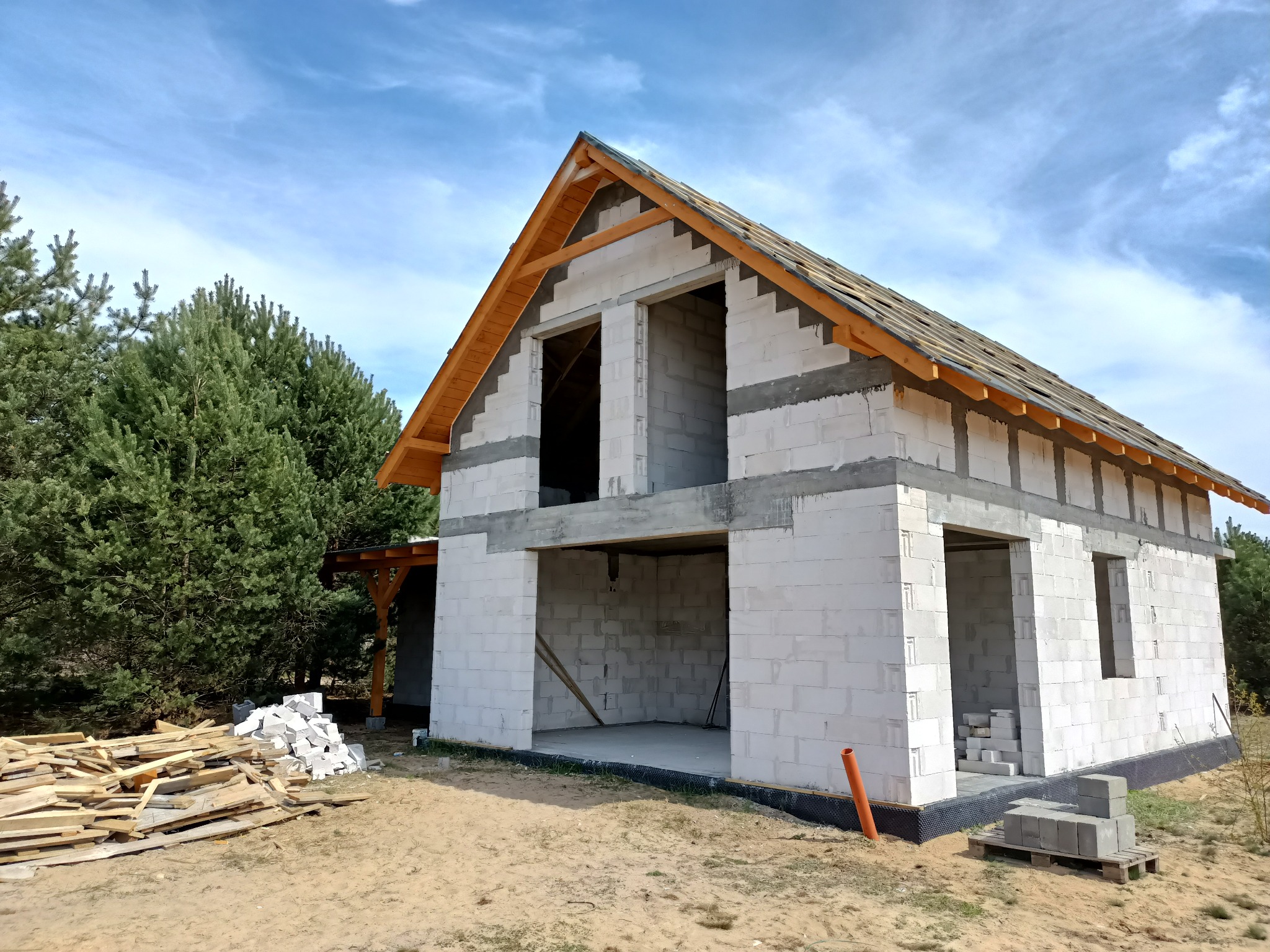 Surowy stan budowy domu jednorodzinnego z białych bloczków, widoczne drewniane elementy konstrukcji dachu, stosy bloczków i desek na zewnątrz, błękitne niebo w tle.