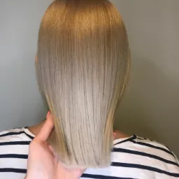 Tył głowy z prostymi, blond włosami w odcieniach od ciemnego blondu u nasady do jasnego na końcach, trzymanymi przez dłoń z pomalowanymi na pomarańczowo paznokciami, na tle gładkiej ściany i osoby...