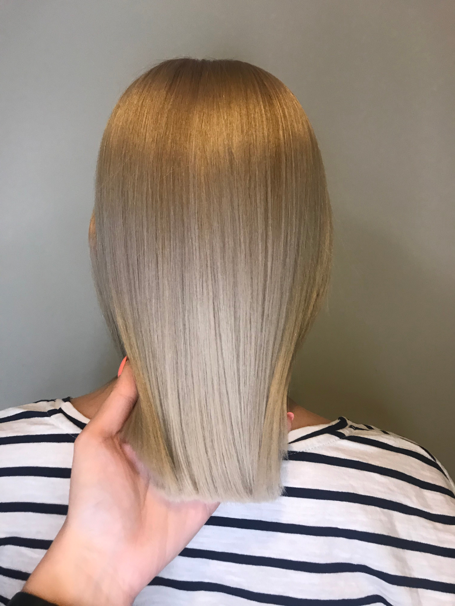 Tył głowy z prostymi, blond włosami w odcieniach od ciemnego blondu u nasady do jasnego na końcach, trzymanymi przez dłoń z pomalowanymi na pomarańczowo paznokciami, na tle gładkiej ściany i osoby...