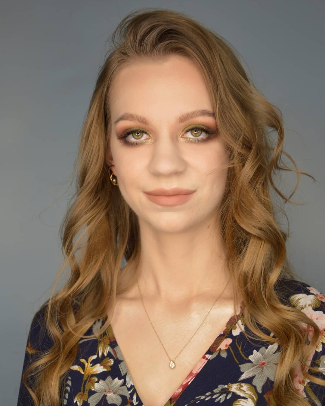 Portret kobiety z delikatnym, złotym cieniem na powiekach, podkreślonymi brwiami i ustami w kolorze nude, w kwiecistej sukience i z drobnym złotym naszyjnikiem.