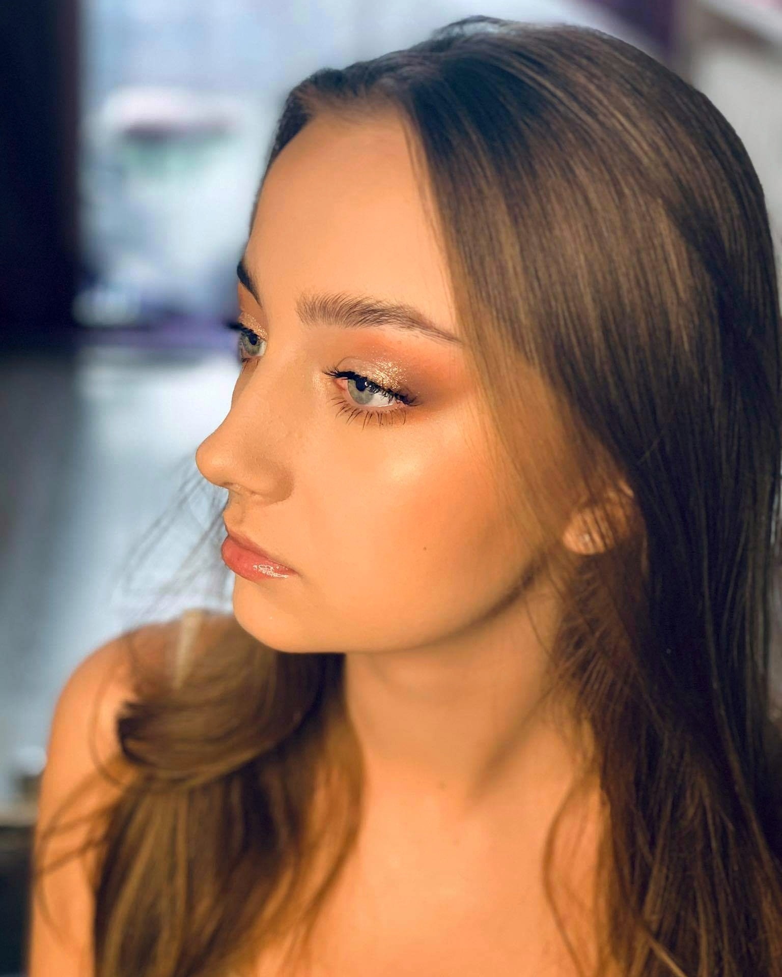 Portret z profilu młodej kobiety z delikatnym, rozświetlonym makijażem oczu w odcieniach złota i brązu, błyszczącymi ustami i subtelnym konturowaniem twarzy, na rozmytym tle.