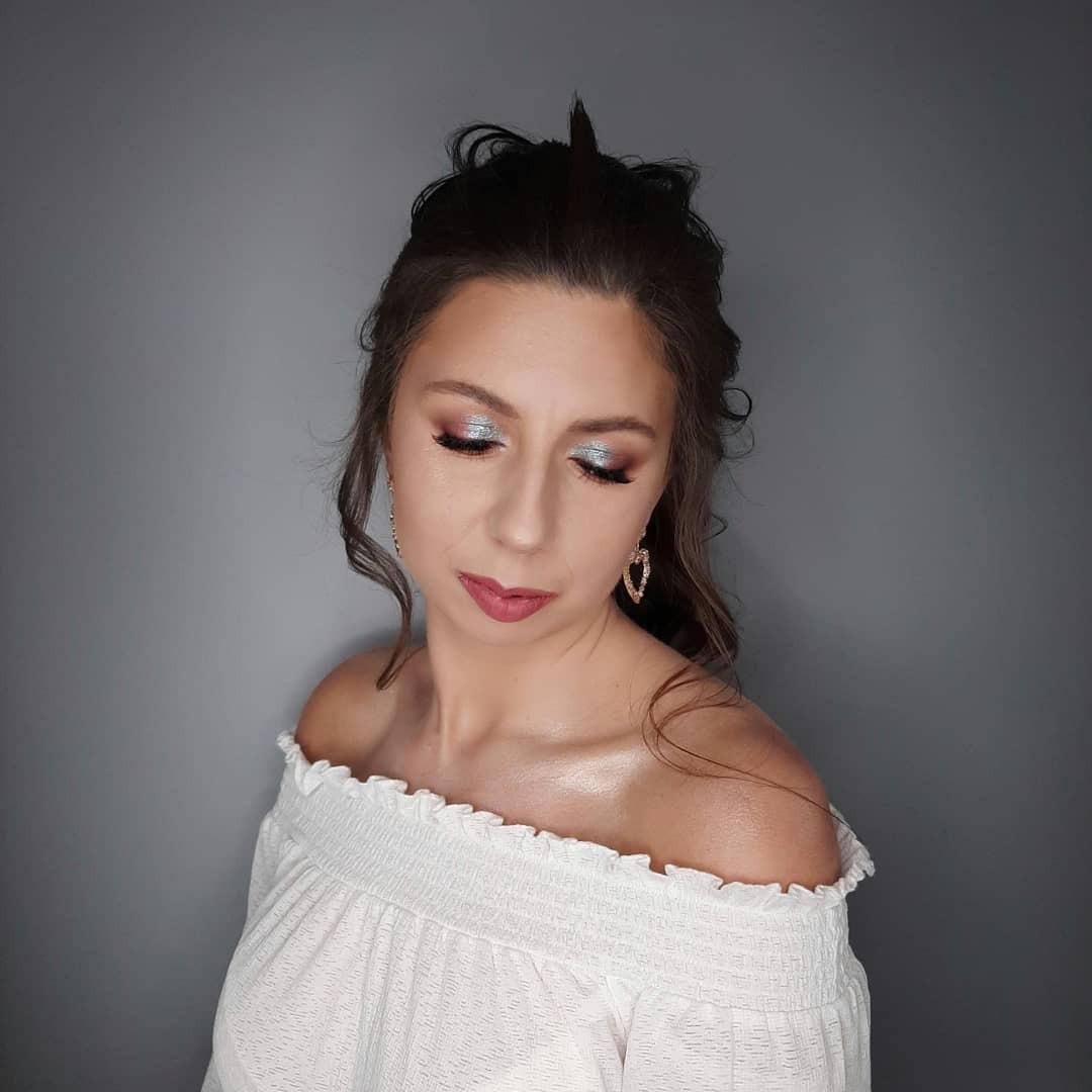 Portret kobiety z upiętymi włosami, srebrzystym cieniem na powiekach i delikatnym różem na ustach, pozującej w białej bluzce z odkrytymi ramionami na tle szarej ściany.
