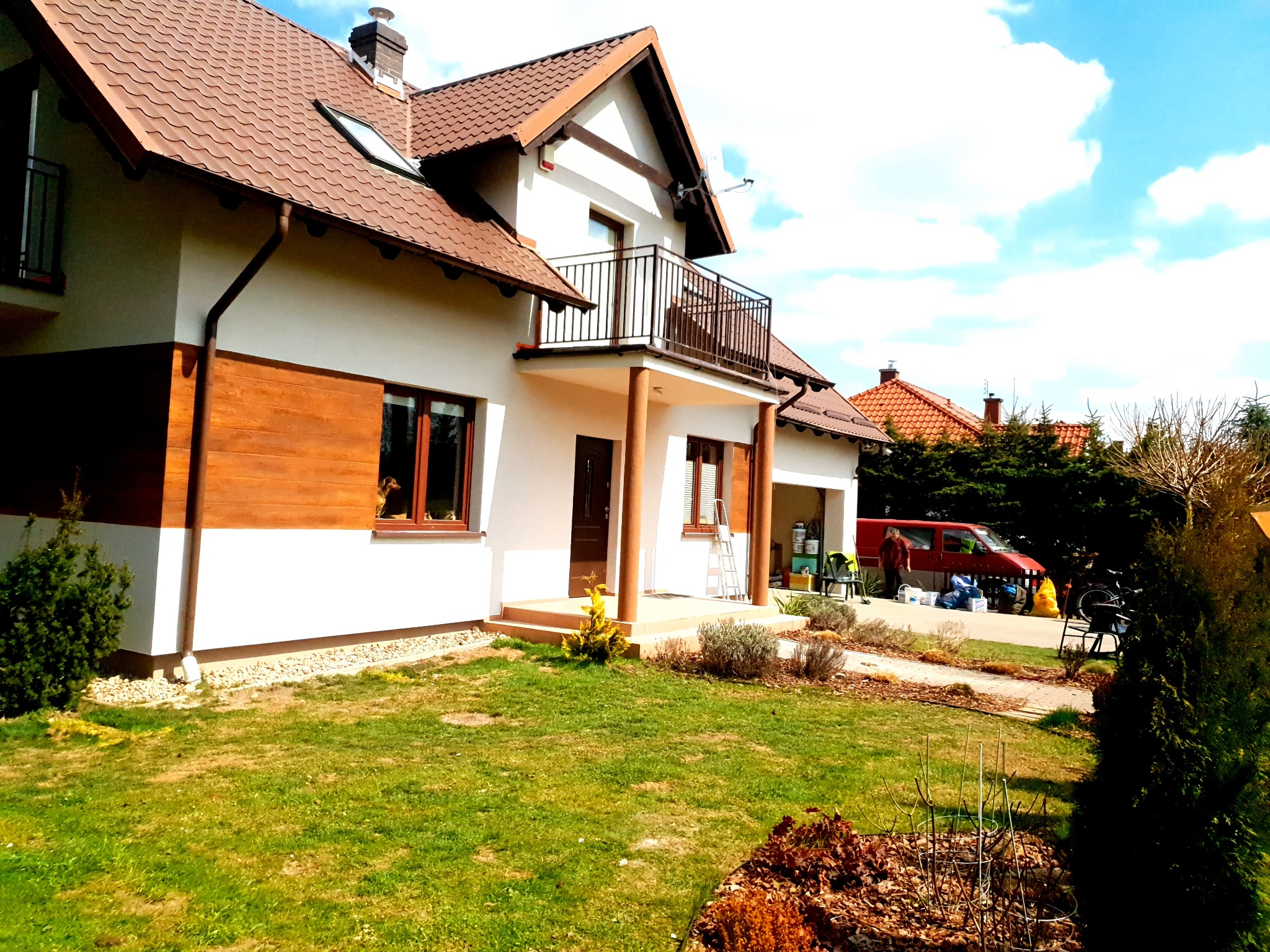 Dwukondygnacyjny dom jednorodzinny z brązowym dachem i częściowo drewnianą elewacją, widoczny z perspektywy ogrodu w słoneczny dzień; na pierwszym planie trawnik, w tle czerwony van i garaż.