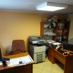 Wnętrze biura z biurkami, komputerami, drukarką wielofunkcyjną i regałem z segregatorami, oświetlone lampą sufitową.