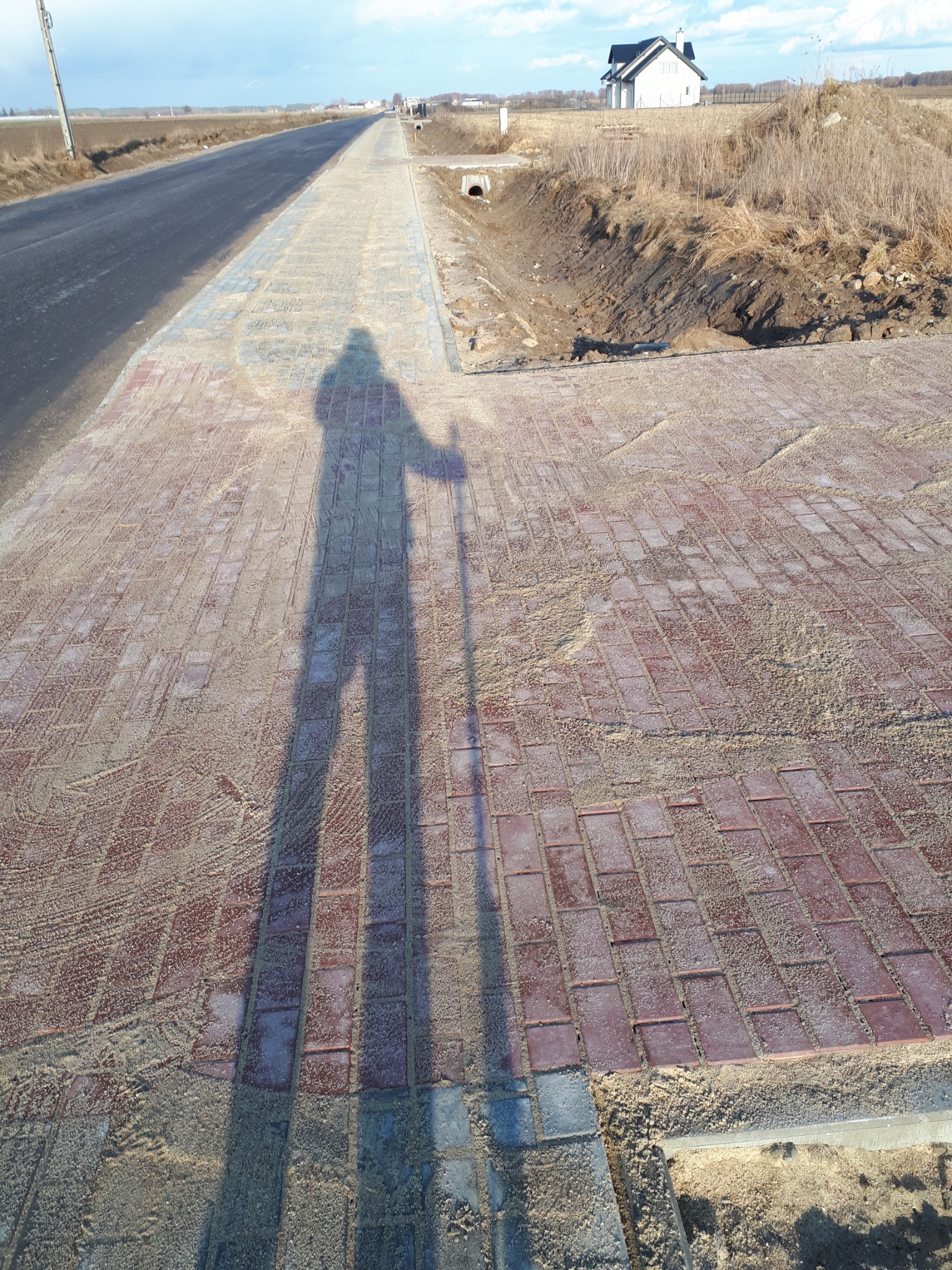 Czerwona kostka brukowa ułożona wzdłuż drogi, pokryta piaskiem, cień postaci z laską w słoneczny dzień.