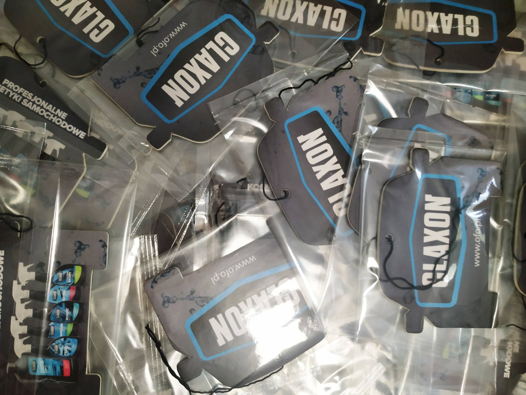 Stos zapakowanych w folię zawieszek zapachowych do samochodu marki Claxon z niebieskimi obwódkami i napisem na szarym tle, widoczny adres strony www.ofo.pl